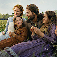 A family of four in old-fashioned clothing sits close together in a grassy field, smiling at each other on a sunny day surrounded by trees and nature, creating a warm, peaceful, rural atmosphere.