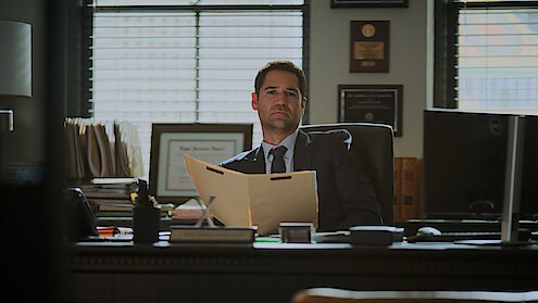 Man in a suit sits at a desk in an office, holding an open folder. Behind him are shelves with files, awards, and plaques, with sunlight streaming through window blinds.