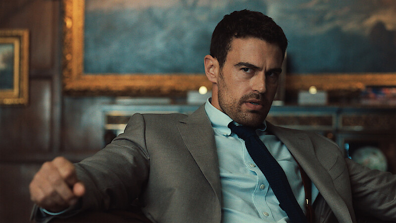 Man in a suit sitting in a formal office, looking serious, with a painting and wood-paneled walls in the background, giving an intense expression.