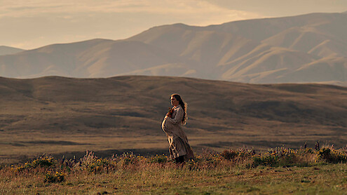 Pregnant woman in a long dress stands in a grassy field with wildflowers, set against a backdrop of rolling hills and mountains during golden hour, creating a serene and natural outdoor scene.