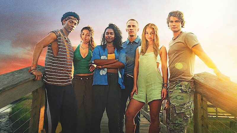 Six young adults stand together on a wooden boardwalk at sunset, with open fields and dramatic skies in the background, posing confidently in casual summer clothing.