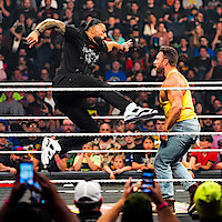 Two wrestlers in a crowded arena; one jumps with a knee strike toward the other inside a wrestling ring, surrounded by ropes, with fans watching and capturing the action on their phones in the background.