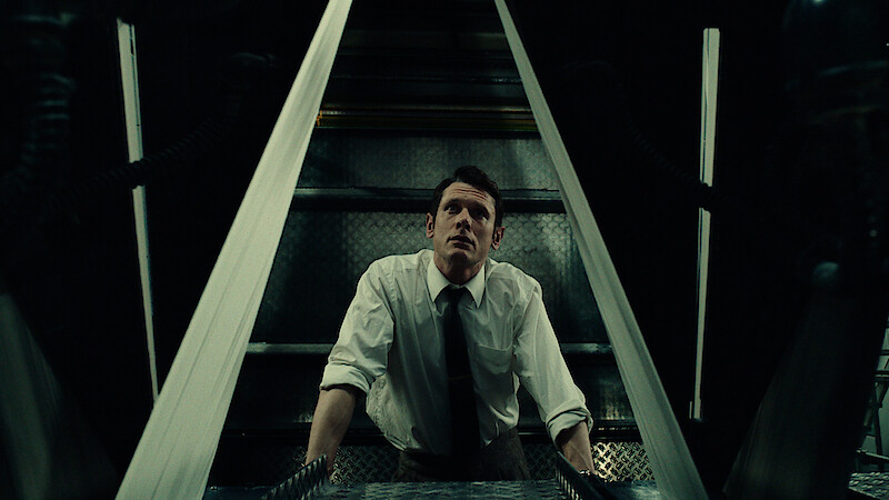 Man in white shirt and tie looks upward in a dimly lit industrial or mechanical setting, framed by metallic structures, suggesting a tense or suspenseful atmosphere.
