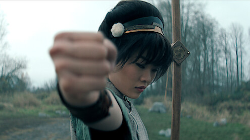 Miyako as Toph in traditional attire strikes a martial arts pose outdoors, focusing forward with a determined expression, holding a staff, with a backdrop of trees and cloudy sky in a natural, open setting.