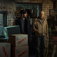 Three people stand in a dimly lit, cluttered basement or storage room with brick walls and stacked boxes. The mood appears tense or serious, and the setting looks old and industrial.