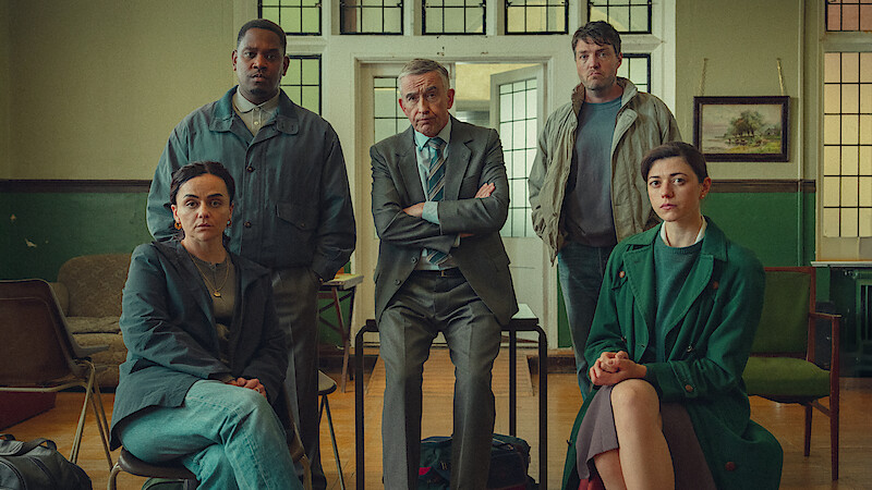 Five adults, three seated and two standing, pose in a serious group setting inside a classroom or office with green walls, wooden floor, chairs, and a painting on the wall, suggesting a meeting or discussion environment.