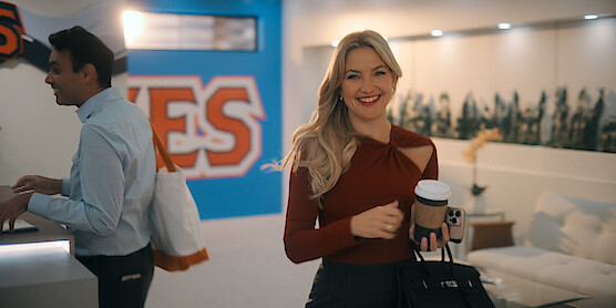 Smiling woman holding coffee and a bag stands in a modern, brightly lit office with a man in the background, wall art reading "YES", and contemporary furniture in the lounge area.