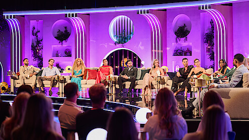 A group of people sit on a brightly lit stage set with purple lighting and art deco design, participating in a panel discussion or reality TV reunion, while an audience watches from the foreground.