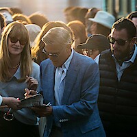 A man in a blue suit signs autographs in a crowded outdoor setting at sunset. People around him wear sunglasses and casual clothing, creating an excited and lively atmosphere.