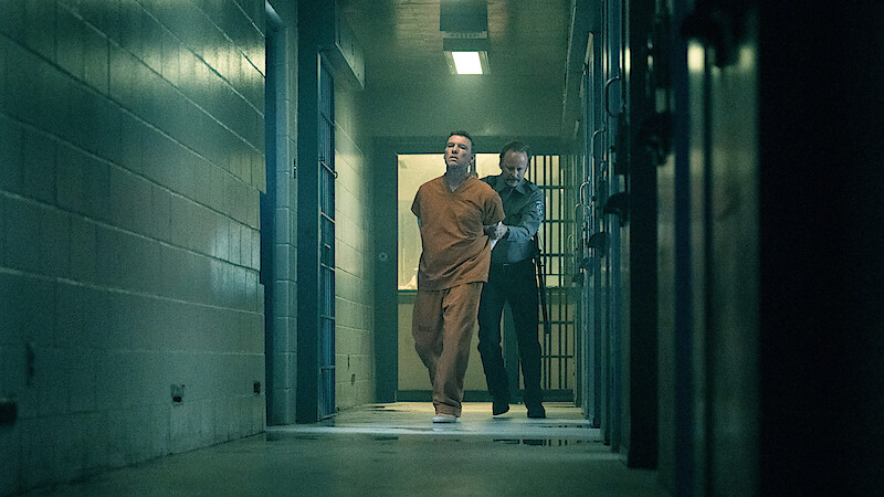 A man in an orange prison jumpsuit is being escorted by a guard down a dimly lit hallway in a jail or prison, with barred cells and concrete walls visible.