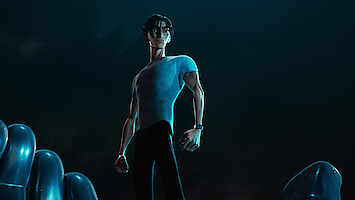 Ken Sato (Christopher Sean) wears a tight gray T-shirt and stands against a black background.