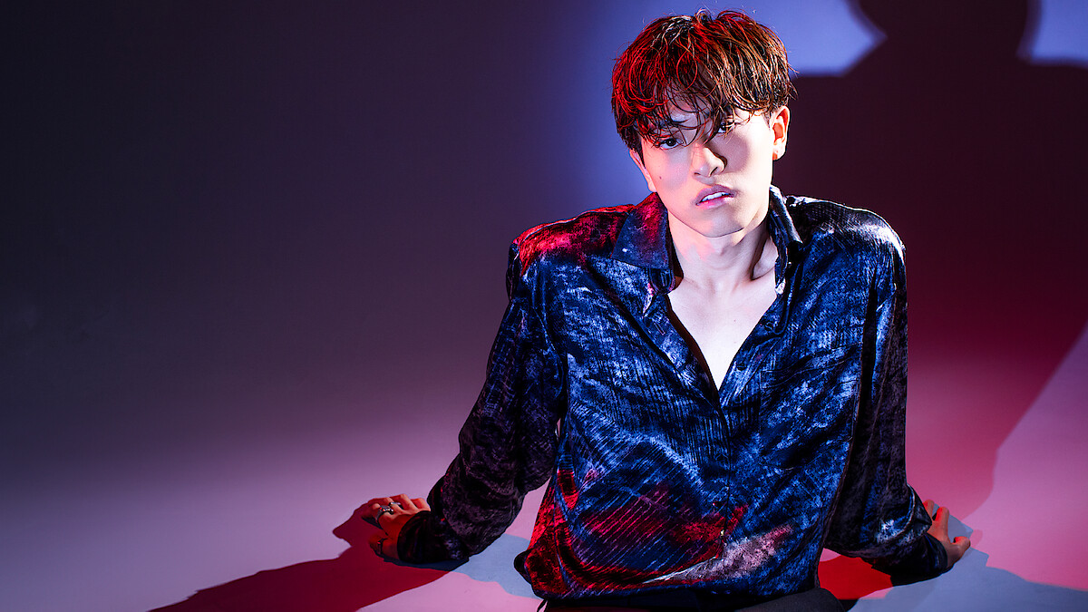 Alan Chikin Chow with wet hair sits on the floor in a dimly lit studio with dramatic red and blue lighting, wearing a shiny dark shirt. Their serious expression and pose cast deep shadows on the wall behind them.