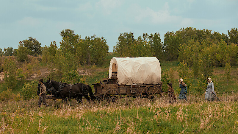 A group of people with horses pulls a covered wagon through a grassy field, surrounded by green trees under a partly cloudy sky, evoking a 19th-century pioneer or settler scene in nature.