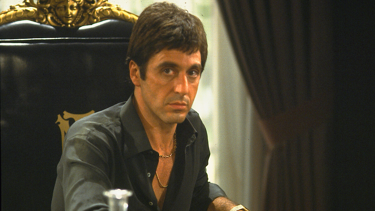 A serious man with short dark hair is seated at a grand, ornate black chair in a dimly lit room with dark curtains and a decorative background, wearing a black shirt and gold necklace.