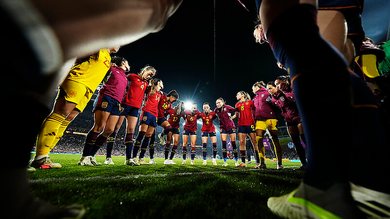 A women's soccer team huddles together on a grass field at night under stadium lights, wearing matching uniforms, preparing for a match with an atmosphere of unity and focus.