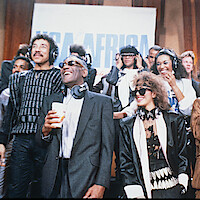Group of musicians wearing headphones in a lively recording studio, smiling and singing together in front of a USA for Africa banner during a charity event.