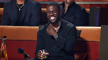 Man in black shirt sitting on a couch at an event, laughing, holding a drink, with a microphone and bottles on the table in front of him, and other people seated in the background.