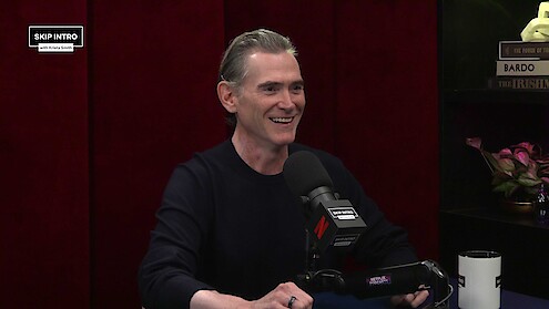 A smiling man sits in a podcast studio with a microphone, dark red curtains, and shelves with books and flowers behind him. The mood is relaxed and conversational, suggesting a friendly interview setting.