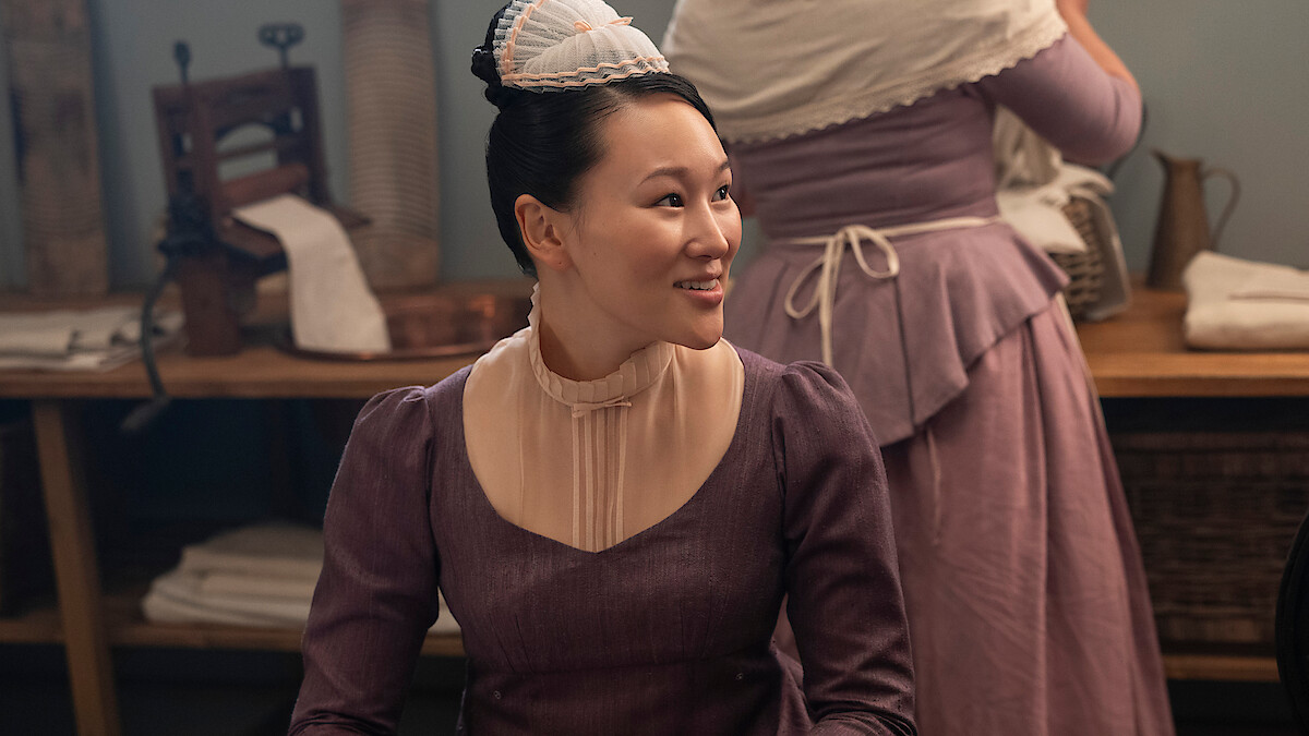 A woman in a purple historical dress and cap sits smiling in a rustic room with wooden tables, linens, and vintage laundry tools, while another similarly dressed woman works in the background.