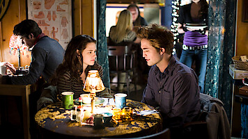 A young woman and man sit at a small round table with drinks and a lamp between them in a cozy, warmly-lit café, talking closely while other people sit and walk in the background.