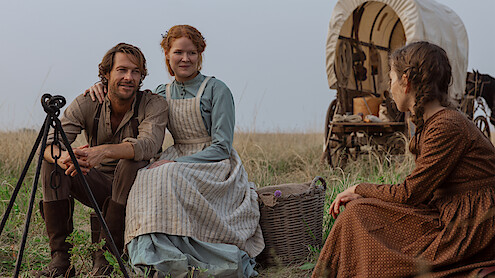 Three people in pioneer-era clothing sit in a grassy field near a covered wagon; two are sitting together and smiling at a third woman, with camping supplies visible, evoking a historical, rural outdoor setting.