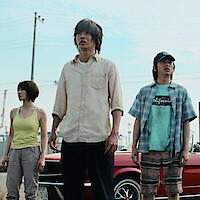 Three young adults stand in front of a red car outdoors, looking ahead with serious expressions on their faces. The setting includes visible power lines, utility poles, and industrial structures in the background.