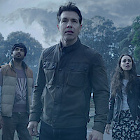 Three people stand outdoors, looking up with concerned expressions. Behind them are tall trees and a cloudy, stormy sky, suggesting an intense or dramatic situation in a natural environment.