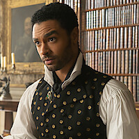 A man in Regency-era clothing sits in a richly decorated library with bookshelves and paintings, while a servant stands in the background near a window and candle holder.