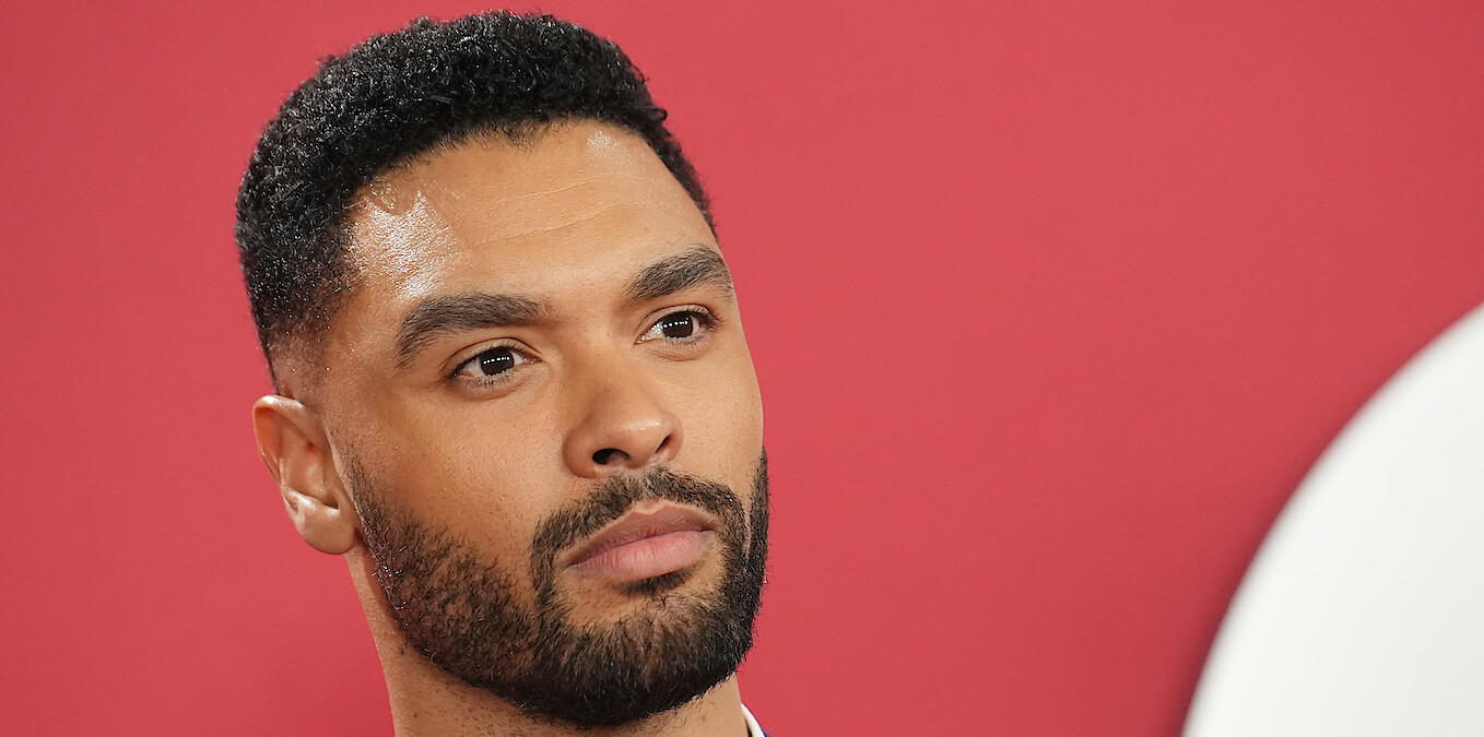  A man with short curly hair and a beard stands against a solid pink background, looking thoughtful and composed, suggesting a formal or professional event atmosphere.