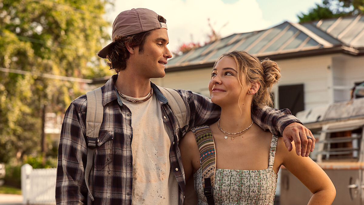 A young man and woman outdoors, smiling at each other. The man wears a cap and plaid shirt, the woman wears a sundress. They are close together with his arm around her, in a sunny, suburban neighborhood with trees and houses.