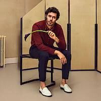 Adam Brody está sentado em um banco segurando uma flor copo-de-leite preta