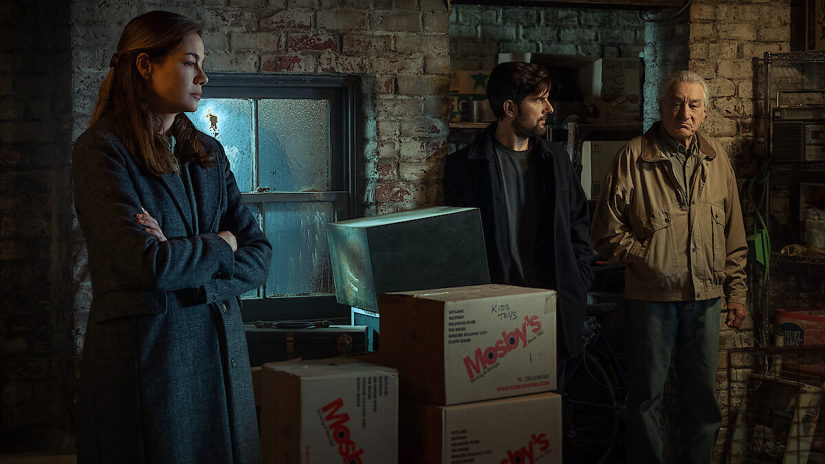 Three people stand in a dimly lit, cluttered basement or storage room with brick walls and stacked boxes. The mood appears tense or serious, and the setting looks old and industrial.