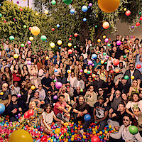 Large group of people gathered outdoors under trees, posing together while colorful balloons float around them, creating a festive, cheerful atmosphere in daylight.