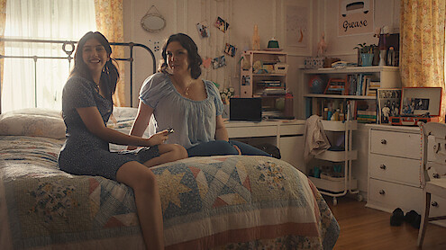 Two women sitting on a bed in a cozy, sunlit bedroom with bookshelves, photos, and personal items in the background, creating a warm and inviting atmosphere.