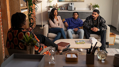 Three people sit on a couch in a modern, warmly lit office, facing another person in a chair, suggesting a therapy or counseling session. The room has plants, shelves, and a coffee table with magazines.