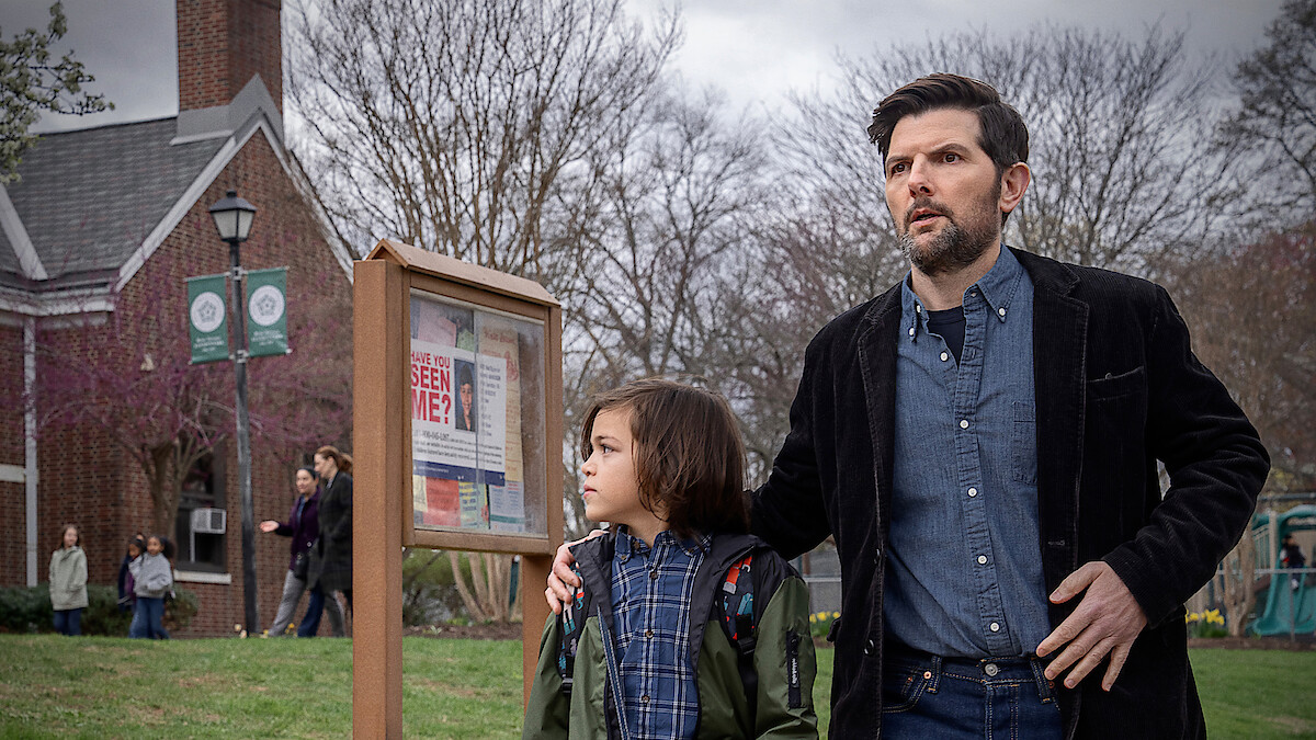 A man and child stand outside near a community notice board with a "Have You Seen Me?" poster. They look alert or concerned. The setting is a grassy area by a brick building with people and leafless trees in the background.