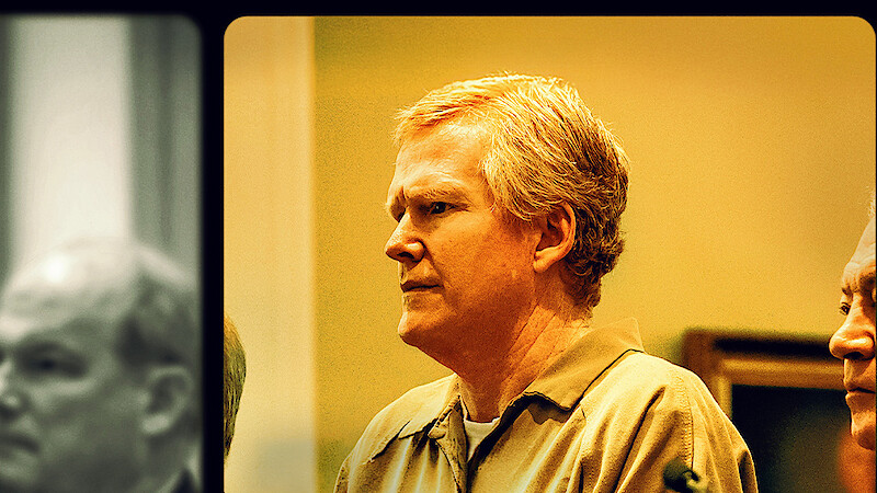 Middle-aged man with light hair in a tan prison uniform stands in a courtroom setting, with another man beside him and blurred figures in the background, all under warm indoor lighting.