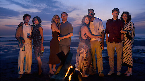 Group of friends standing together on a beach at sunset near a campfire, dressed in casual evening clothes, some holding drinks, ocean and colorful sky in the background, creating a relaxed and warm atmosphere.
