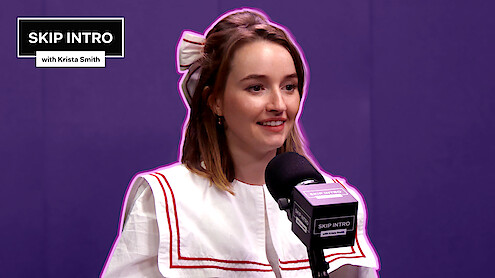 Kaitlyn Dever, smiling warmly, is seated in front of a microphone.