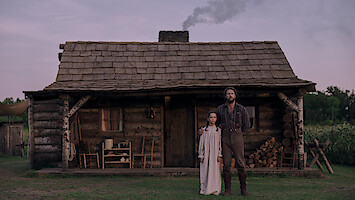 A man and a girl stand in front of a rustic log cabin with a wooden porch, chairs, stacked firewood, and smoke rising from the chimney. The setting is rural, with grass and cornfields under a purple evening sky.