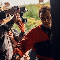 Man in red and black jacket pushing through crowd of photographers outdoors, with cameras pointed at him, surrounded by people and greenery in the background.