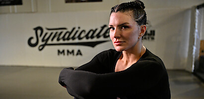 Gina Carano with braided hair in athletic wear sits with arms crossed in an MMA gym, "Syndicate MMA" visible on the wall behind her, suggesting a focused training environment.