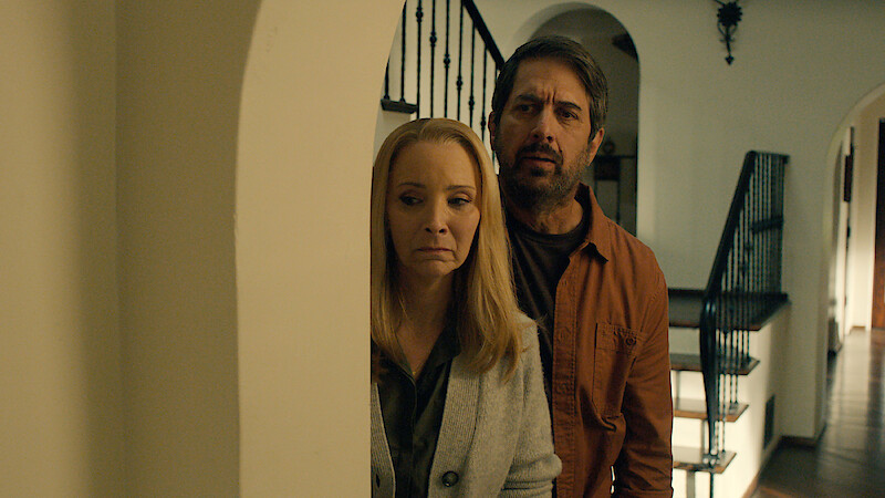 Ray Romano as Paul and Lisa Kudrow as Lydia hide behind a doorway with worried expressions in Season 1 of 'No Good Deed'