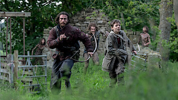 Two people, one man and one boy, run through a grassy, wooded area with stone ruins, both holding bows, while several figures follow behind them in the background. The scene is tense and outdoors in a natural environment.
