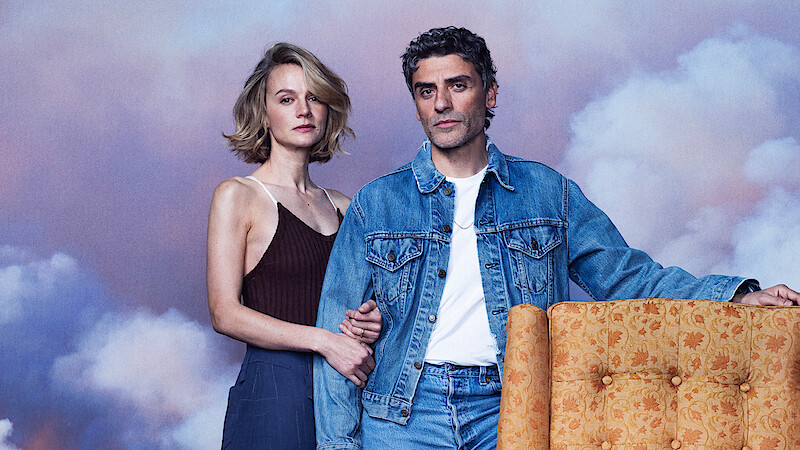 A man in a denim jacket and a woman in a sleeveless top stand together indoors by an orange armchair, with a soft, cloudy sky backdrop creating a calm, dreamy atmosphere.