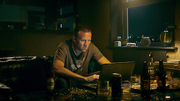 A man sits in a dimly lit, cluttered room at night, focusing intently on his laptop. The environment feels tense and isolated, with bottles and plates scattered around a table, suggesting loneliness or stress.