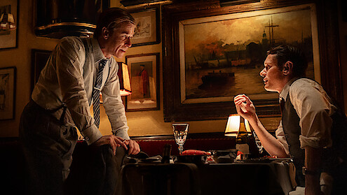 Two men in dress shirts converse seriously at a softly lit restaurant table, surrounded by framed paintings and classic decor, with wine and glasses on the table, creating an intimate, vintage atmosphere.