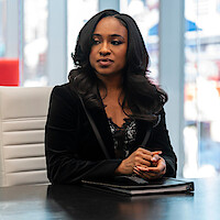 A woman with long hair sits at a conference table in a modern office, wearing a black blazer and lace top, hands folded over a closed folder, with large windows and blurred cityscape in the background.