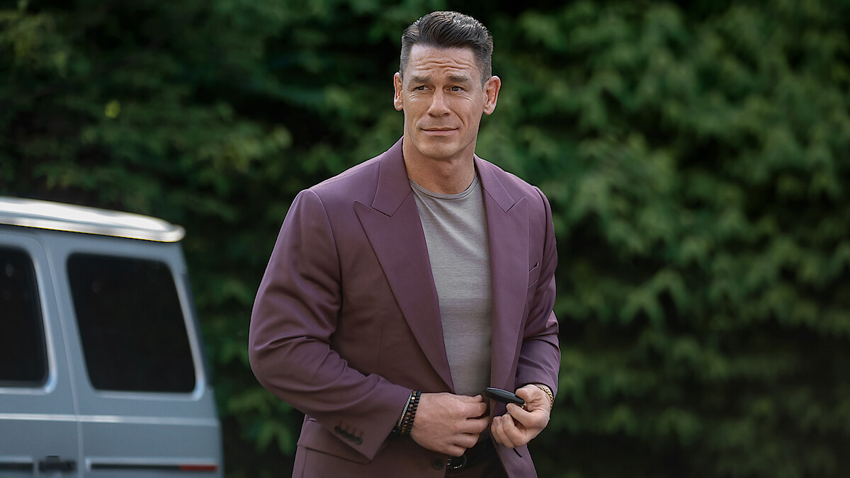 Man in a mauve suit jacket and gray shirt stands outdoors near a silver van, with a background of dense green foliage, holding an object and looking slightly to the side.
