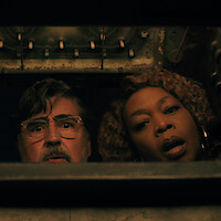A man and a woman with surprised expressions look down into a dimly lit compartment or container, suggesting curiosity or discovery in an industrial or mechanical setting in 'The Boroughs.'